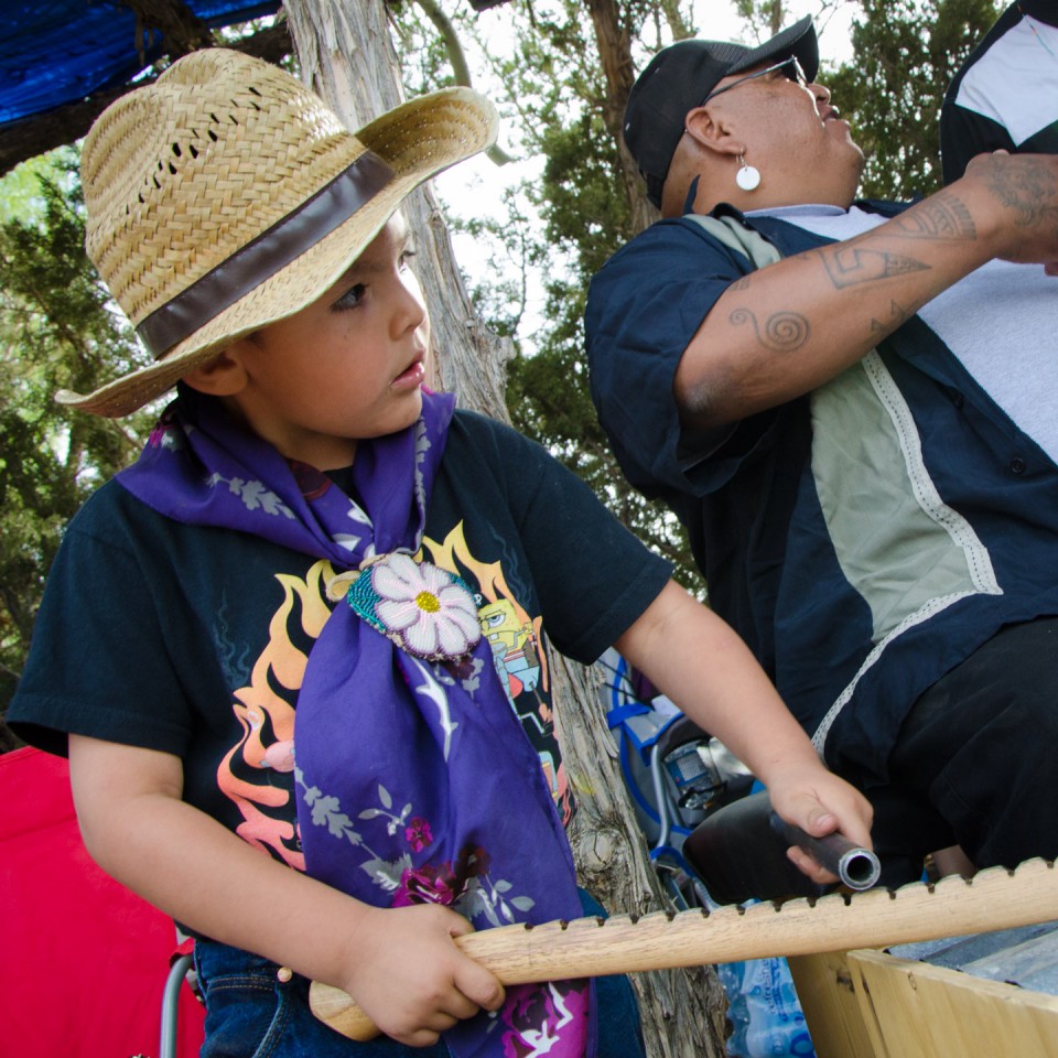 A well-dressed youth makes Bear Dance music, sitting alongside more experienced Ute singers.